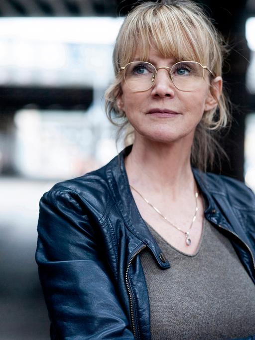 Eine Frau blickt nach links oben. Sie trägt eine Brille und ein graues Oberteil, dazu eine schwarze Jacke. Es ist die Schauspielerin Esther Esche.