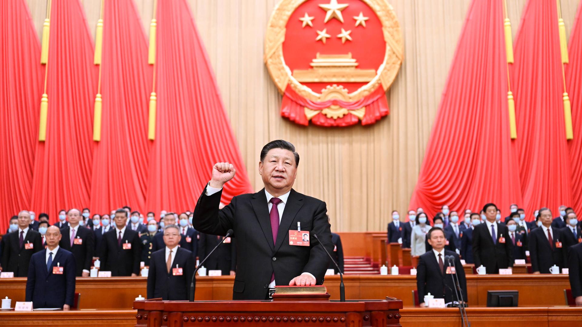 Chinas Präsident steht mit erhobener rechter Faust hinter einem Rednerpult, die andere Hand liegt auf einem Buch. Hinter ihm das Parteiemblem der KPCh, rote Fahnen und viele Volkskongressabgeordnete.