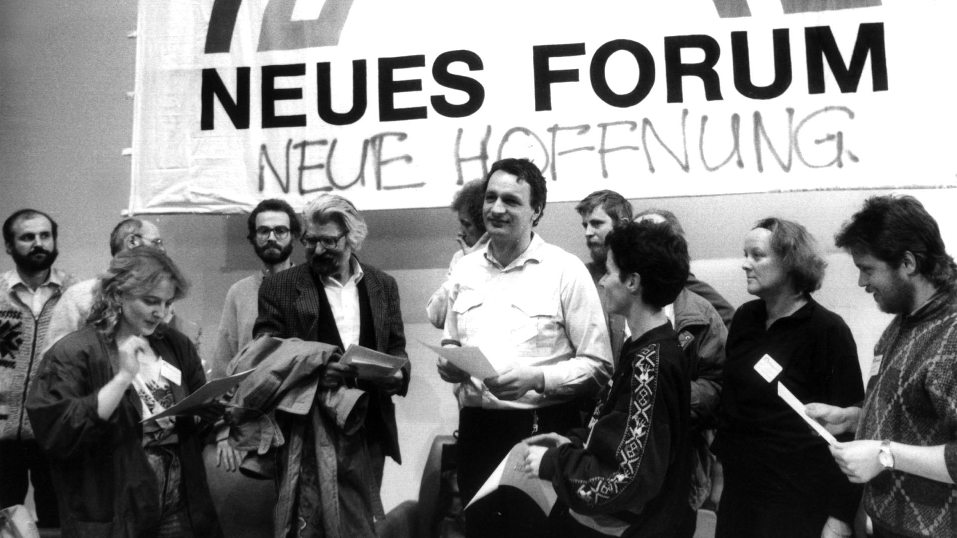 Schwarzweiß-Fotografie von einer Gruppe Menschen, die vor einem Banner mit der Aufschrift "Neues Forum" stehen. Darunter steht geschrieben "Neue Hoffnung". 