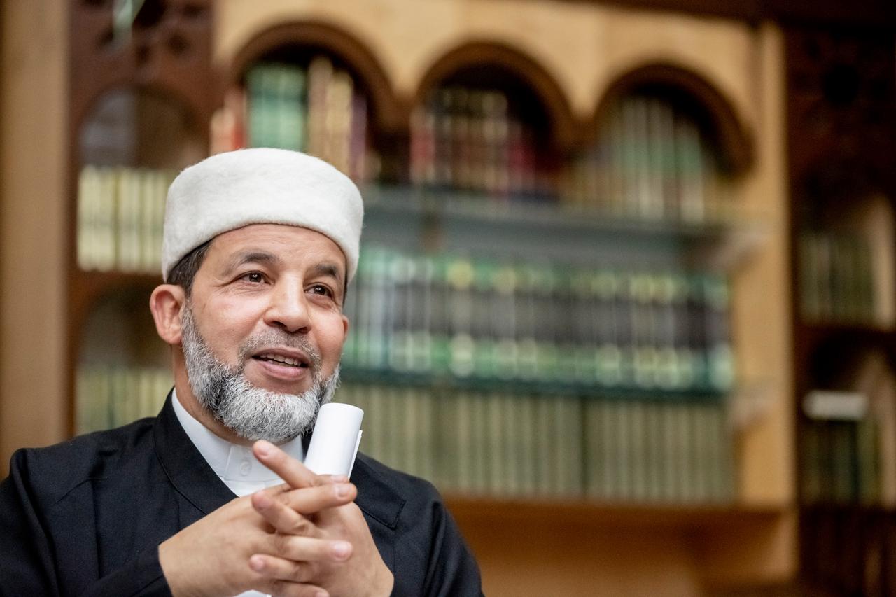 Imam Taha Sabri mit grauem Bart und schwarzer Kleidung. Im Hintergrund ist verschwommen ein Regal mit Büchern zu erkennen.