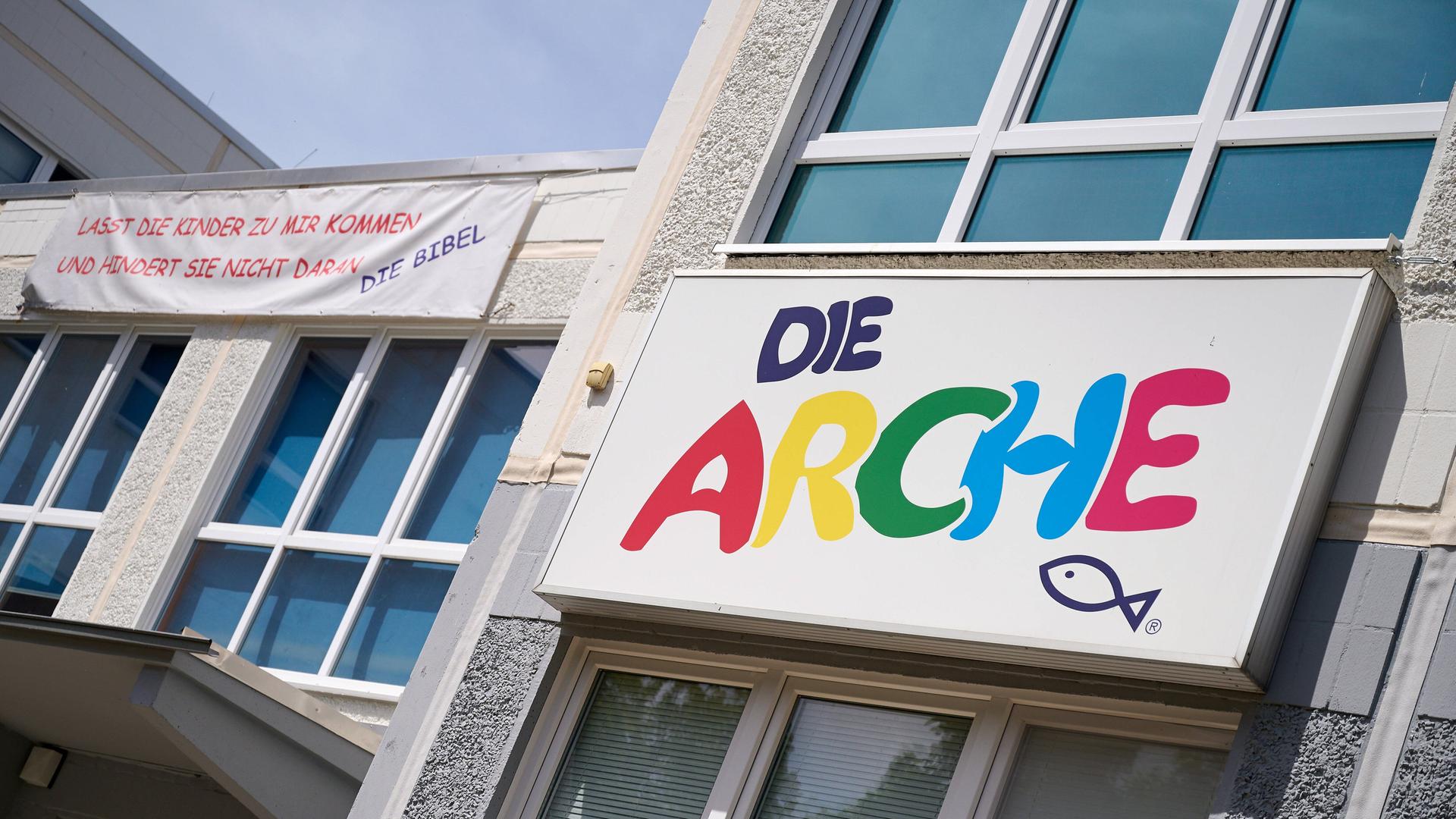 An einer Hauswand hängt ein Schild mit der Aufschrift Die Arche in bunten Buchstaben, darunter ist ein Fisch gemalt. Links vom Schild hängt ein Banner mit der Aufschrift Lasst die Kinder zu mir kommen und hindert sie nicht daran, Die Bibel. An einer Hauswand hängt ein Schild mit der Aufschrift Die Arche in bunten Buchstaben, darunter ist ein Fisch gemalt. Links vom Schild hängt ein Banner mit der Aufschrift Lasst die Kinder zu mir kommen und hindert sie nicht daran, Die Bibel.