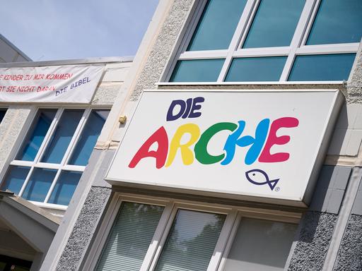 An einer Hauswand hängt ein Schild mit der Aufschrift Die Arche in bunten Buchstaben, darunter ist ein Fisch gemalt. Links vom Schild hängt ein Banner mit der Aufschrift Lasst die Kinder zu mir kommen und hindert sie nicht daran, Die Bibel.