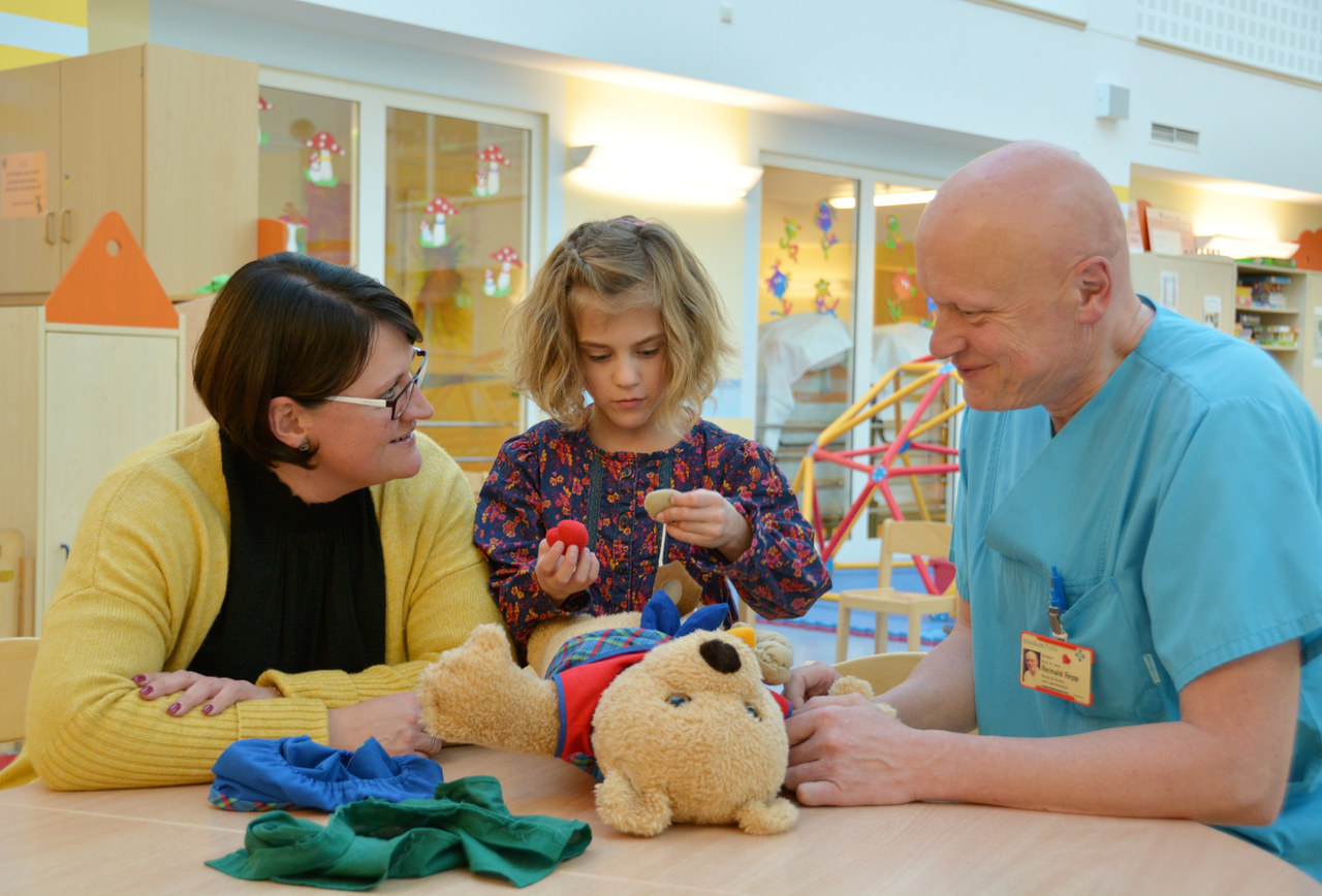 09.11.2018, Hessen, Fulda: Die achtjährige Frieda "versorgt" im Spielraum in der Klinik für Kinder- und Jugendmedizin im Klinikum Fulda einen Teddybär, links ihre Mutter Yvonne, rechts Reinald Repp, Direktor der Klinik. 09.11.2018, Hessen, Fulda: Die achtjährige Frieda "versorgt" im Spielraum in der Klinik für Kinder- und Jugendmedizin im Klinikum Fulda einen Teddybär, links ihre Mutter Yvonne, rechts Reinald Repp, Direktor der Klinik.