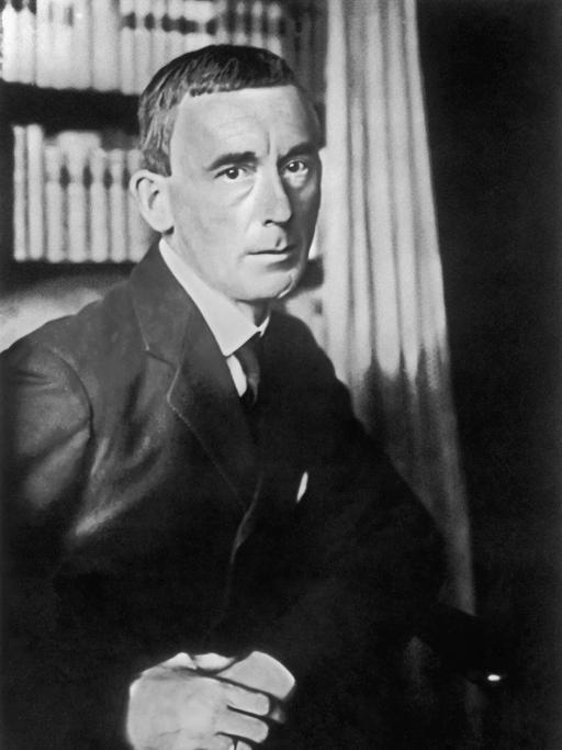 Ein schwarz-weiß Porträt des Schriftstellers Hugo Ball (1886-1927) mit Hemd und Krawatte, hinter ihm ein Bücherregal.