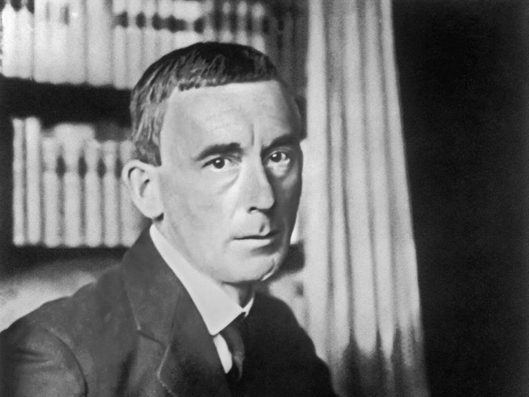 Ein schwarz-weiß Porträt des Schriftstellers Hugo Ball (1886-1927) mit Hemd und Krawatte, hinter ihm ein Bücherregal.