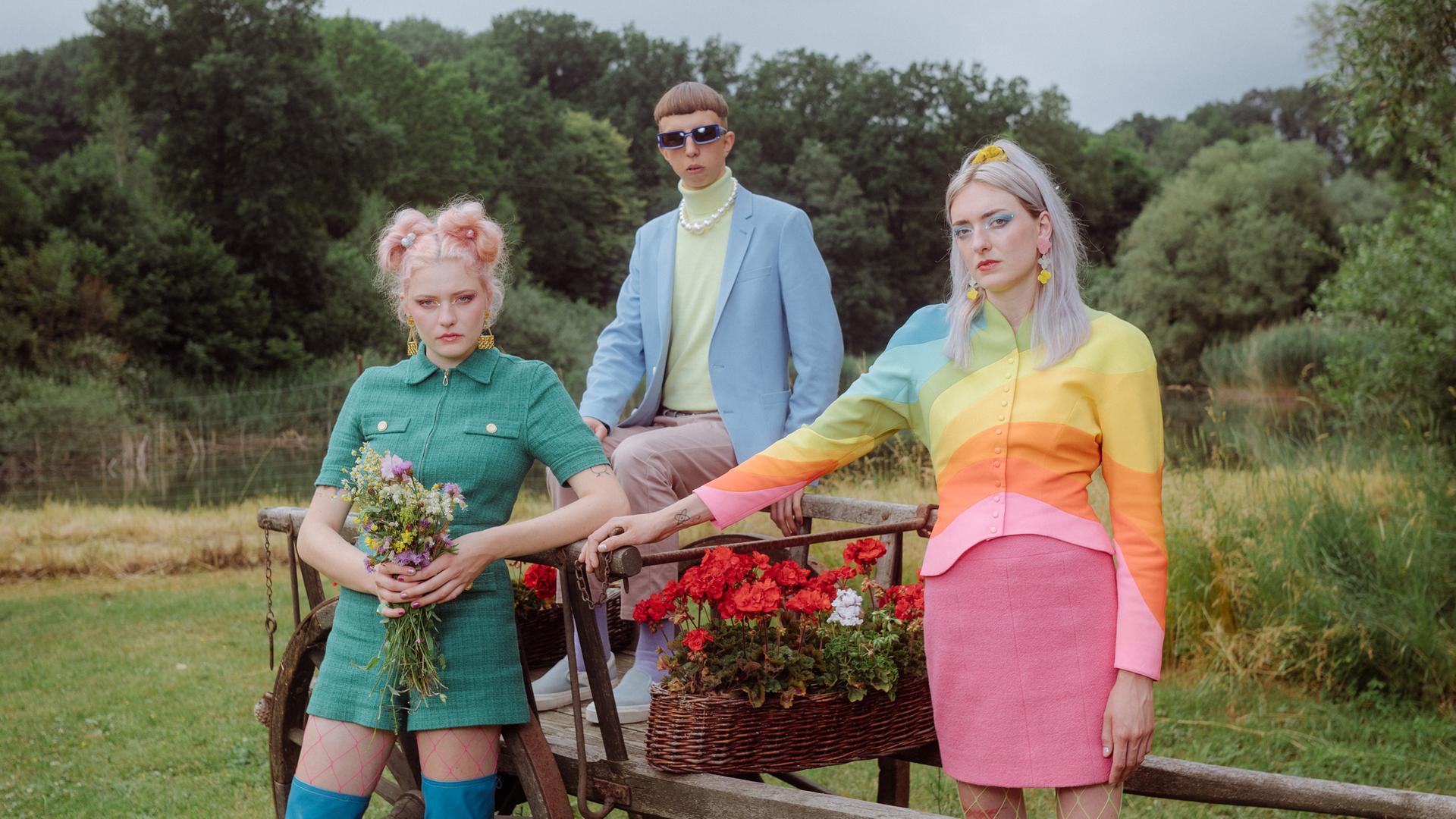 Die dreiköpfige Band Blond, bestehend aus den beiden Schwestern Nina und Lotta Kummer sowie Johann Bonitz posieren auf einer Wiese vor und auf einem blumenbestückten, alten Holzanhänger. Sie tragen poppig-bunte Kleidung. Die dreiköpfige Band Blond, bestehend aus den beiden Schwestern Nina und Lotta Kummer sowie Johann Bonitz posieren auf einer Wiese vor und auf einem blumenbestückten, alten Holzanhänger. Sie tragen poppig-bunte Kleidung.