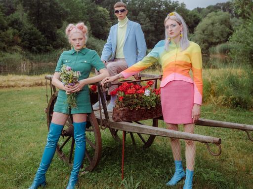 Die dreiköpfige Band Blond, bestehend aus den beiden Schwestern Nina und Lotta Kummer sowie Johann Bonitz posieren auf einer Wiese vor und auf einem blumenbestückten, alten Holzanhänger. Sie tragen poppig-bunte Kleidung.