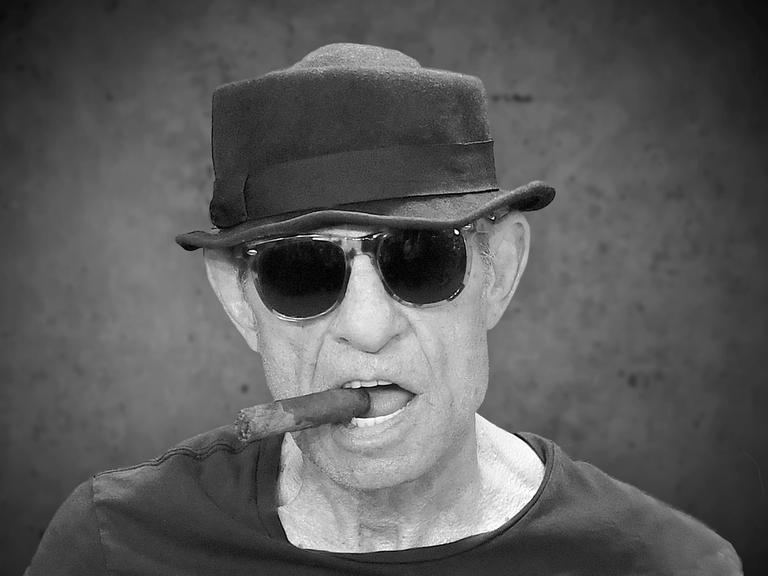 Porträt von Regisseur Klaus Lemke mit einer Zigarre im Mundwinkeln und Hut.