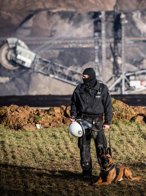 Ein Polizist in voller Montur steht zusammen mit einem angeleinten Hund am Rande des Tagebaus in Lützerath bei Erkelenz. Im Hintergrund ist ein riesiger Bagger im Tagebau zu sehen.