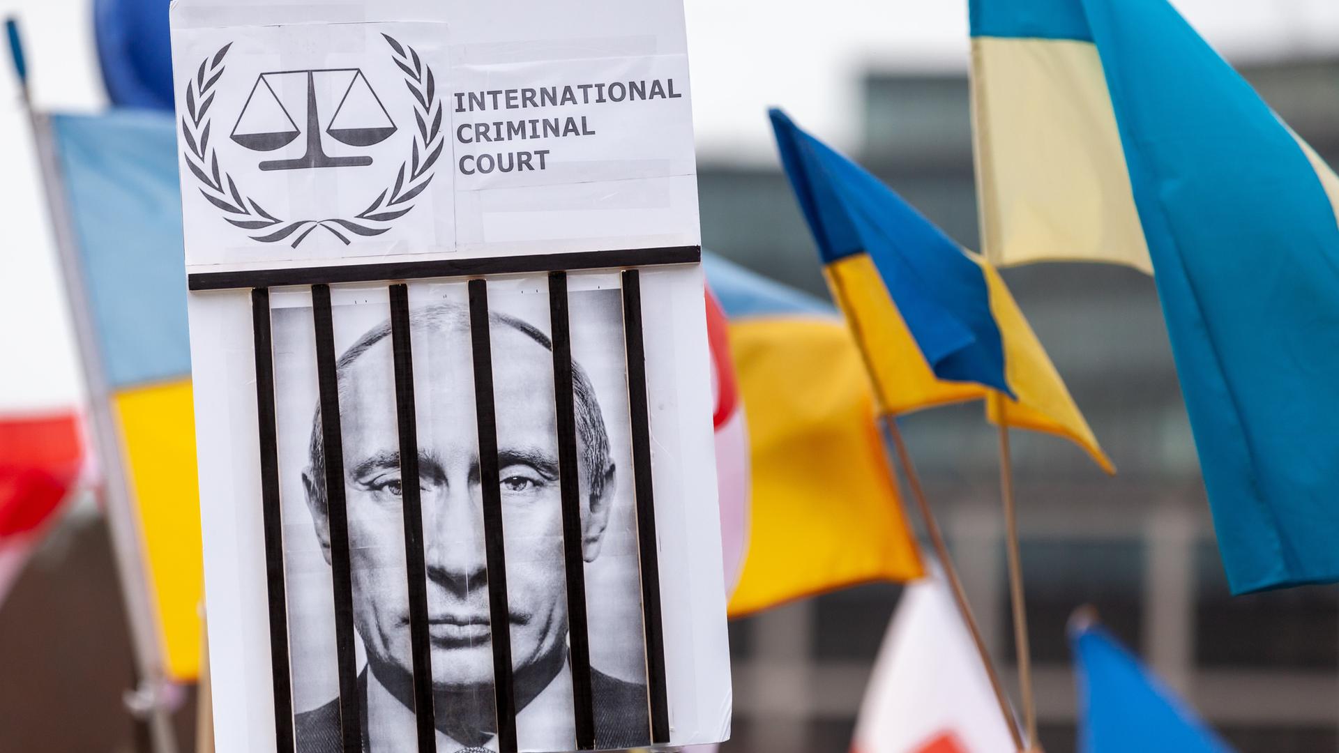 Zwischen Fahnen wird bei einer Demonstration auch ein Plakat hochgehalten, das Russlands Präsident Wladimir Putin hinter Gittern zeigt und den Schriftzug Internationaler Gerichtshof trägt.  
