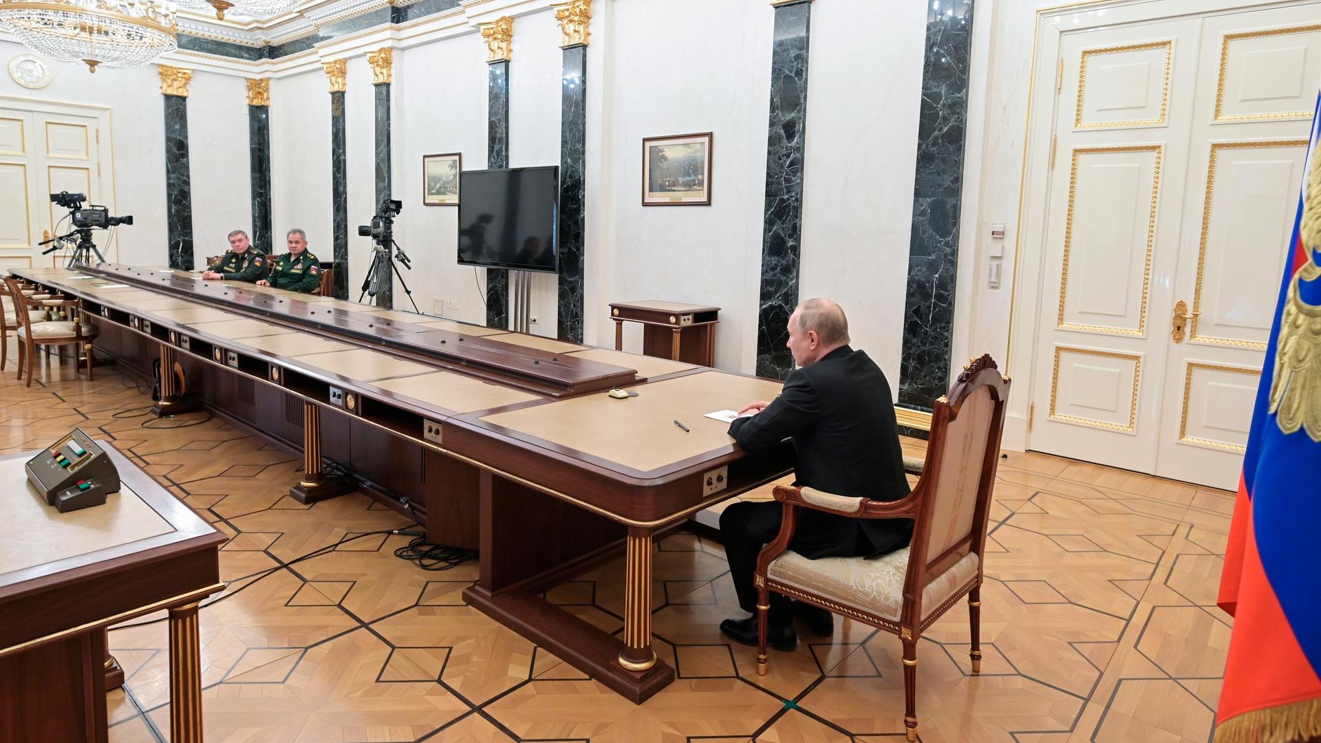 Wladimir Putin sitzt an einem überlangen Tisch im Gespräch mit seinem Verteidigungsminister und einem General der Streitkräfte. 