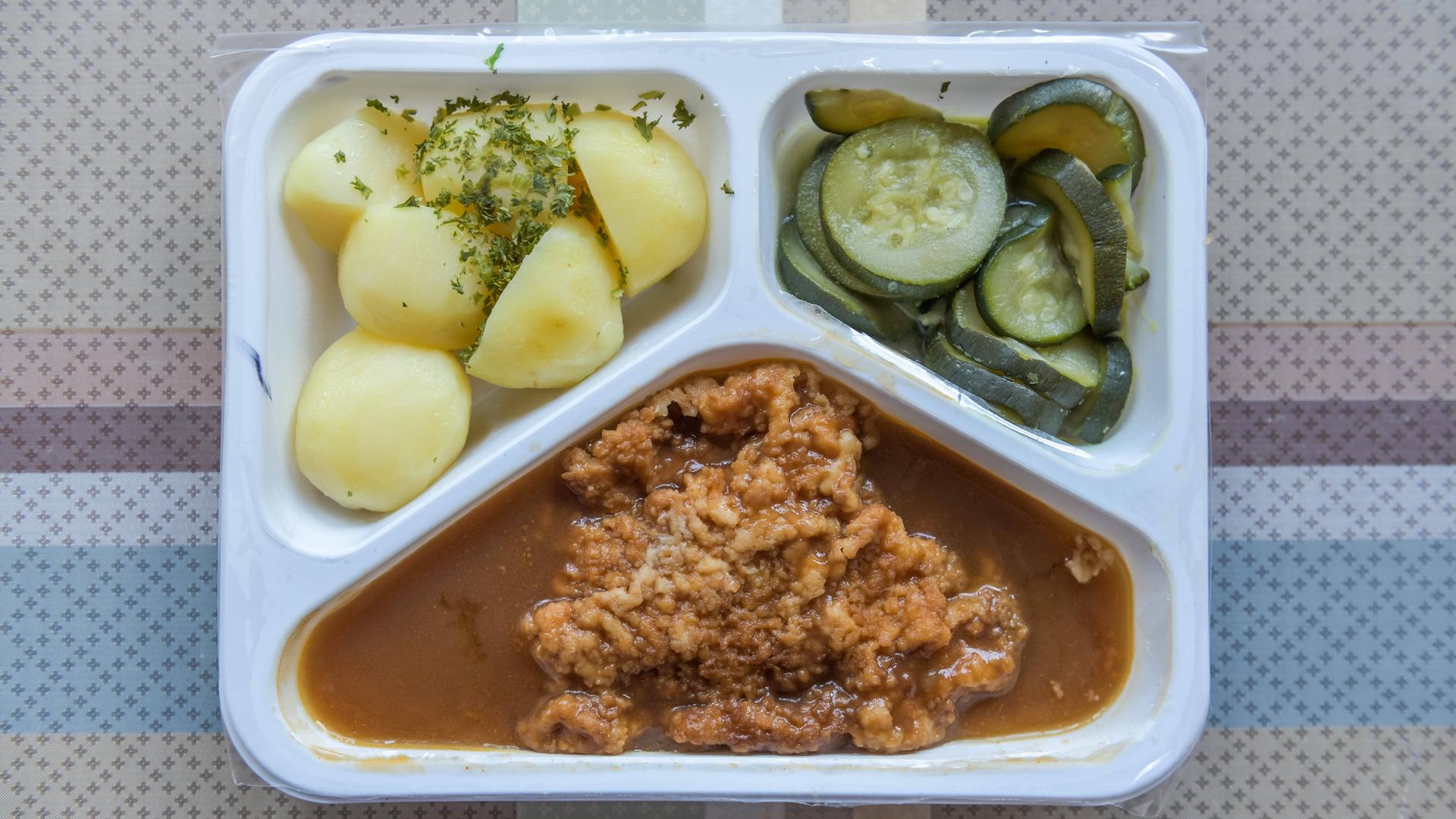 Eine Schale mit drei Teilen steht auf einem Tisch. Sie ist mit Essen gefüllt. In einem Teil sind Kartoffeln. Im zweiten Teil ist ein Schnitzel. Im dritten Teil sind Gurken. Eine Schale mit drei Teilen steht auf einem Tisch. Sie ist mit Essen gefüllt. In einem Teil sind Kartoffeln. Im zweiten Teil ist ein Schnitzel. Im dritten Teil sind Gurken.