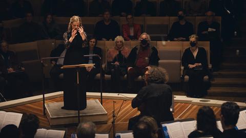 Eine Frau steht vor einem Dirigentenpult, sie gibt mit den Händen Zeichen. Hinter ihr sieht man das Konzertpublikum. Vor ihr steht ein Chor, von den Sängerinnen und Sängern kann man nur die Hinterköpfe erkennen.
