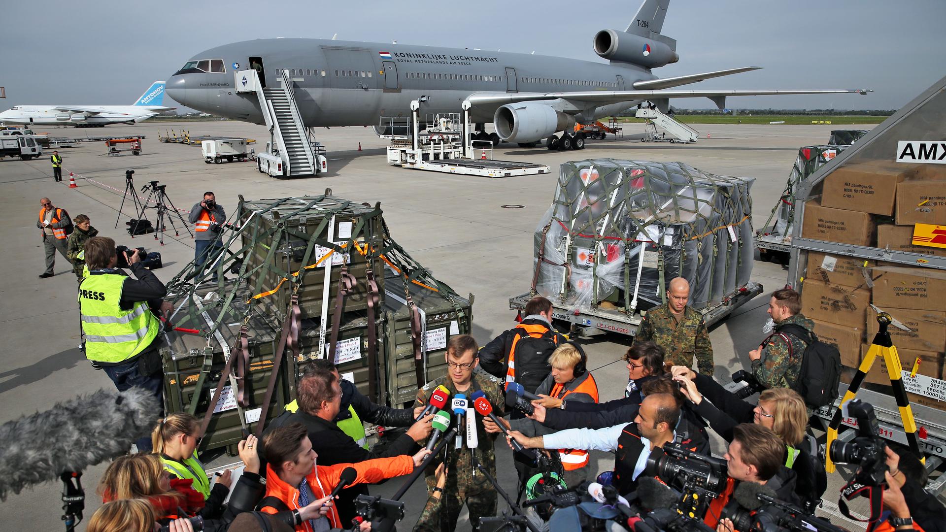 Auf einem Flughafen werden Waffen in ein Transportflugzeug geladen. Im Vordergrund richten zahlreiche Journalistinnen und Journalisten ihre Mikrofone auf einen Sprecher der Bundeswehr. Auf einem Flughafen werden Waffen in ein Transportflugzeug geladen. Im Vordergrund richten zahlreiche Journalistinnen und Journalisten ihre Mikrofone auf einen Sprecher der Bundeswehr.