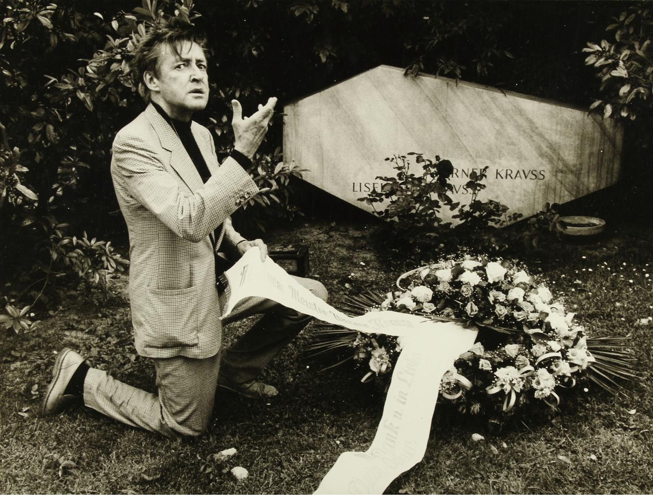 Der Schauspieler Oskar Werner kniet gestikulierend mit einem großen Kranz vor  dem Grab von Burgtheaterschauspieler Werner Krauß am Wiener Zentralfriedhof