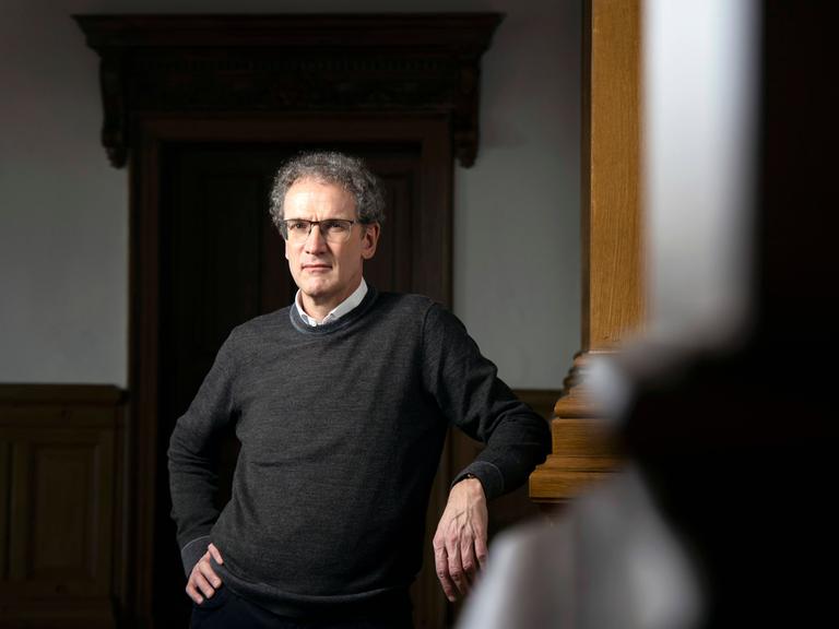 Porträt des Schriftstellers und Literaturwissenschaftlers Dirk Oschmann. Er trägt ein weißes Hemd und einen schwarzen Pullover. Oschmann lehnt an einer hölzernen Säule.