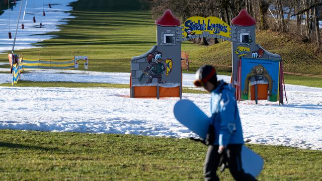 Eine Junge geht mit seinem Snowboard am Draxlhang im Skigebiet Brauneck über eine grüne Wiese. Viele Skigebiete in Bayern leiden aktuell unter Schneemangel.