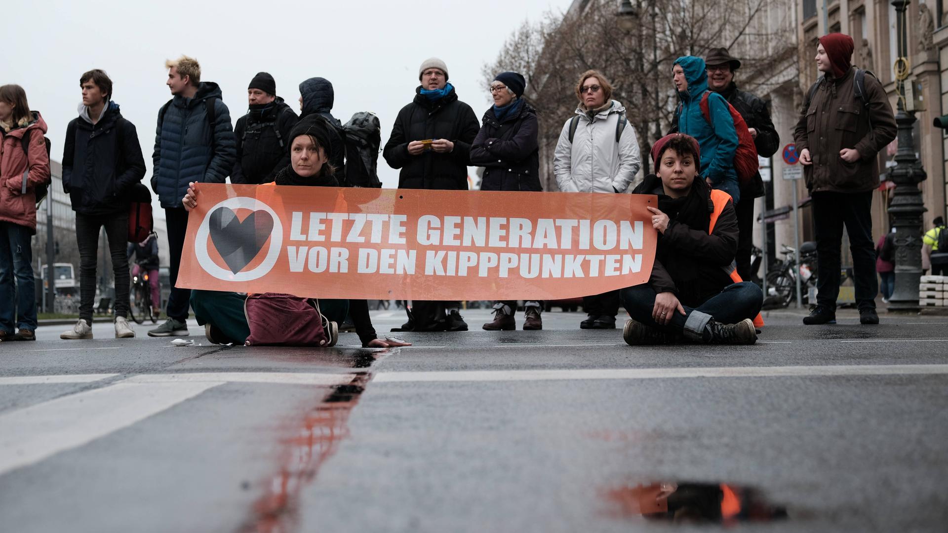 Mehrere Menschen blockieren eine Straße in Berlin mit einem Transparent, auf dem steht: "Letzte Generation vor den Kipppunkten". Mehrere Menschen blockieren eine Straße in Berlin mit einem Transparent, auf dem steht: "Letzte Generation vor den Kipppunkten".