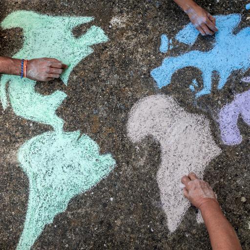 Eine Gruppe von Kindern mal mit bunter Kreide eine Weltkarte auf die Straße. Zu sehen sind nur die Hände.