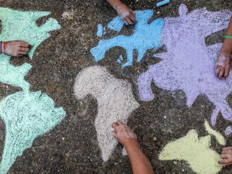 Eine Gruppe von Kindern mal mit bunter Kreide eine Weltkarte auf die Straße. Zu sehen sind nur die Hände.