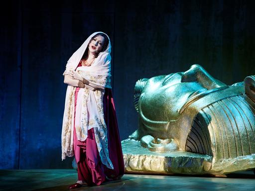 Eine Frau in einem roten Kleid und mit einem hellen Schleier steht vor einem großen goldenen Kopf. Der Kopf sieht wie ein ägyptischer Pharaoh aus. Sie singt und blickt traurig. Es ist die Figur "Aida" aus der Oper von Giuseppe Verdi.