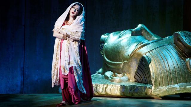 Eine Frau in einem roten Kleid und mit einem hellen Schleier steht vor einem großen goldenen Kopf. Der Kopf sieht wie ein ägyptischer Pharaoh aus. Sie singt und blickt traurig. Es ist die Figur "Aida" aus der Oper von Giuseppe Verdi.