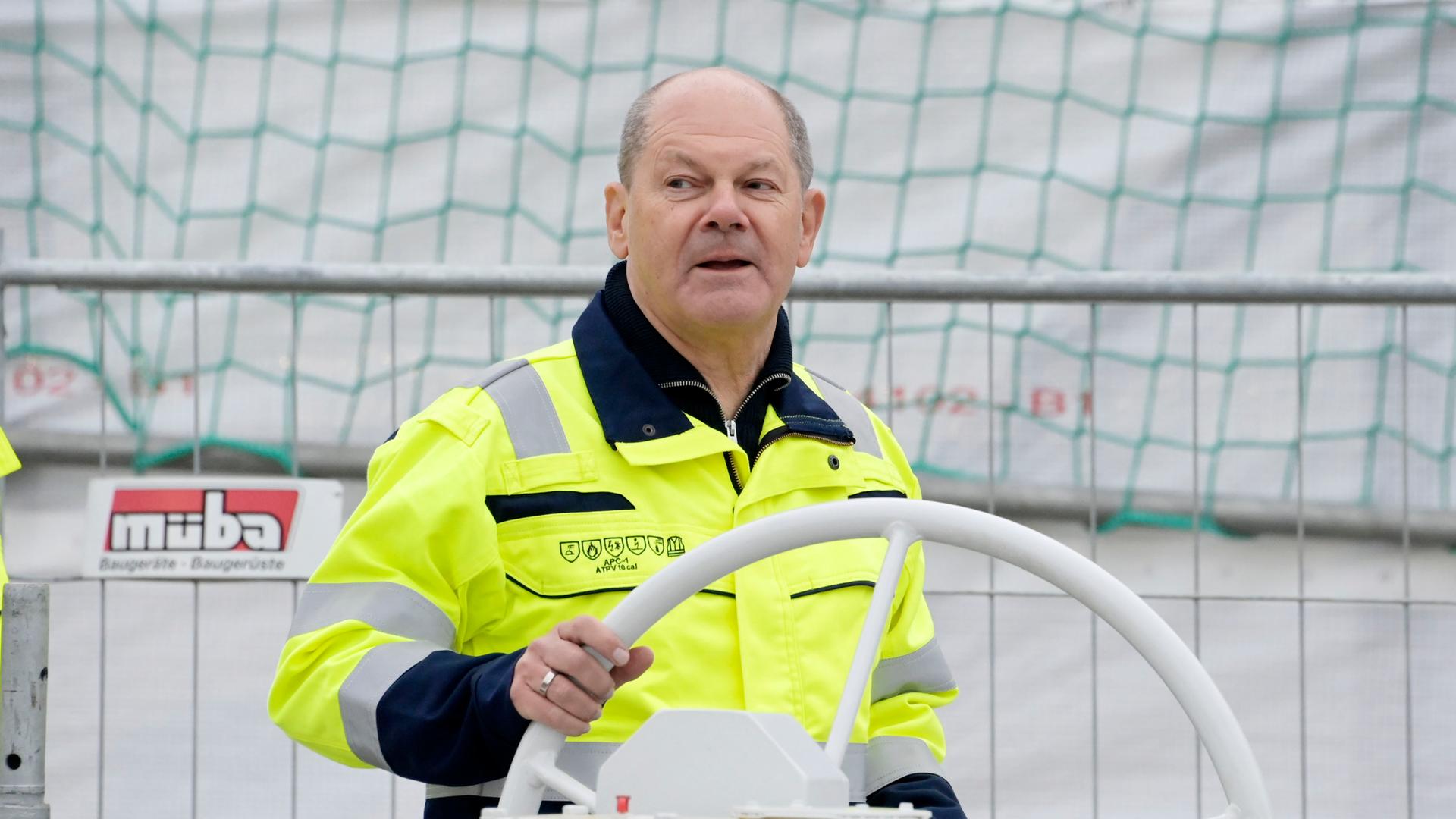 Bundeskanzler Olaf Scholz bei der offiziellen Inbetriebnahme des LNG-Terminals "Deutsche Ostsee" in Lubmin: Er steht hinter einem weißen Steuerrad, trägt eine gelbe Warnweste und blickt zur Seite. Bundeskanzler Olaf Scholz bei der offiziellen Inbetriebnahme des LNG-Terminals "Deutsche Ostsee" in Lubmin: Er steht hinter einem weißen Steuerrad, trägt eine gelbe Warnweste und blickt zur Seite.