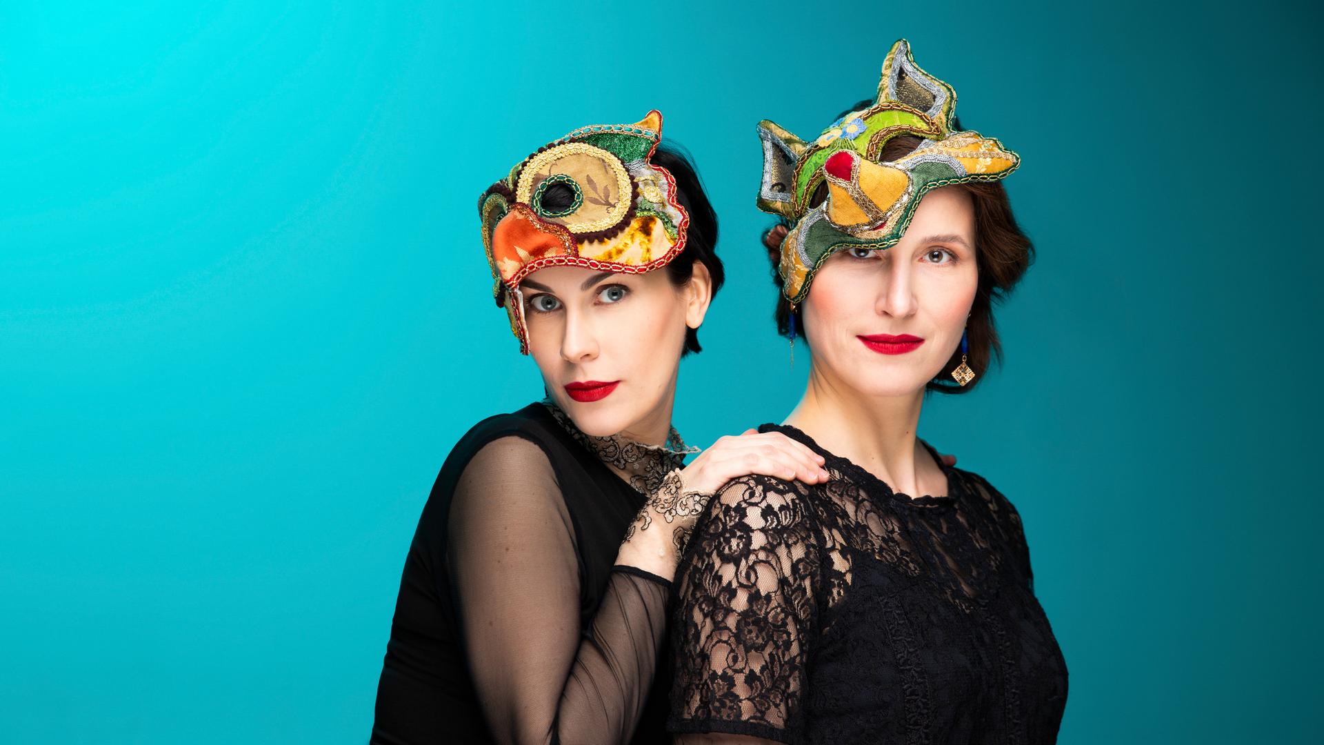 Zwei Frauen stehen nebeneinander vor hellblauem Hintergrund. Sie tragen schwarze Kleidung und auf den Köpfen bunte Masken. Diese kann man sich vors Gesicht ziehen und damit Augen und Nase verdecken. Zwei Frauen stehen nebeneinander vor hellblauem Hintergrund. Sie tragen schwarze Kleidung und auf den Köpfen bunte Masken. Diese kann man sich vors Gesicht ziehen und damit Augen und Nase verdecken.
