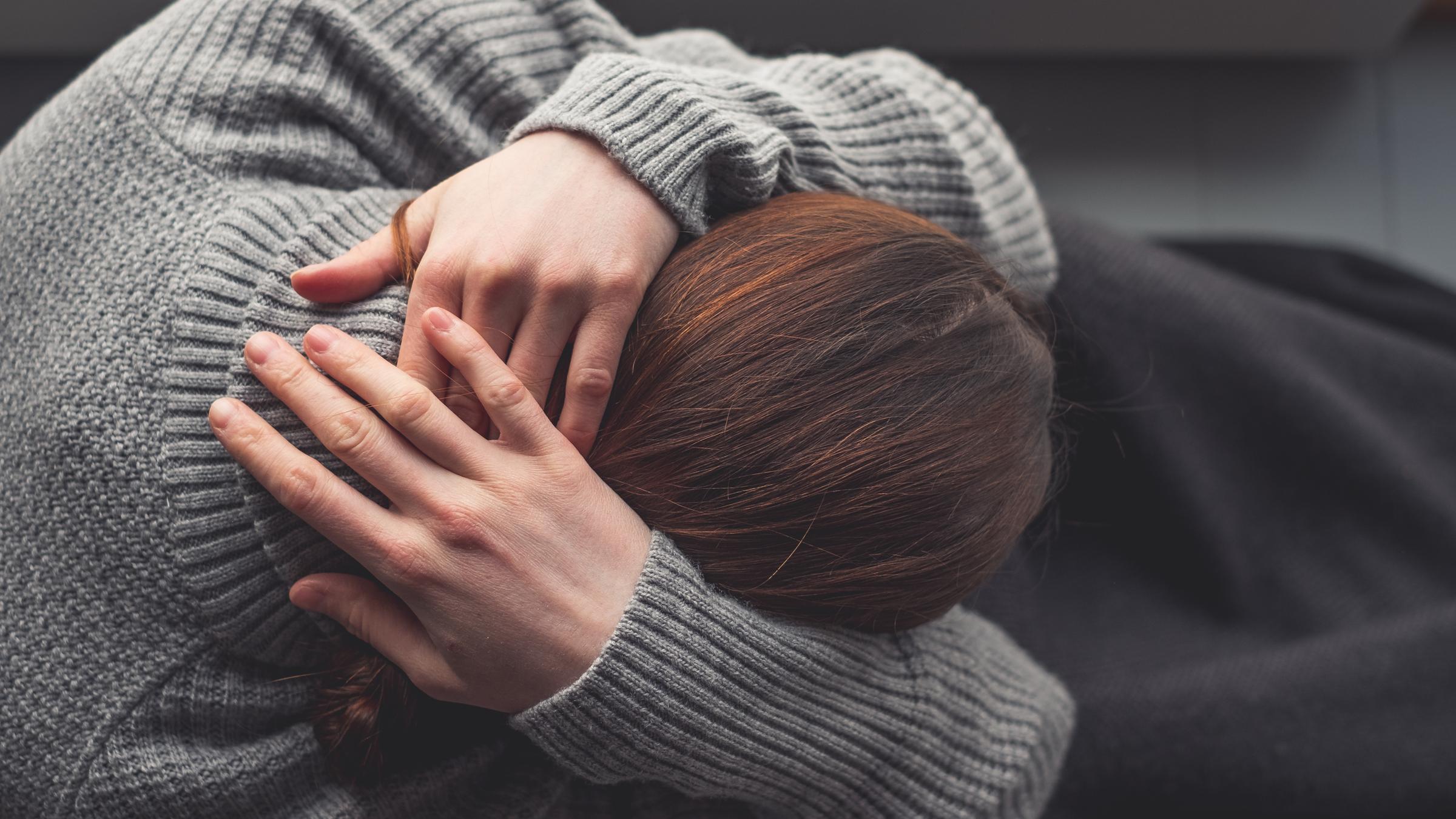 Eine Frau sitzt, die Hände schützend über den Kopf gelegt, an enem Fenster.