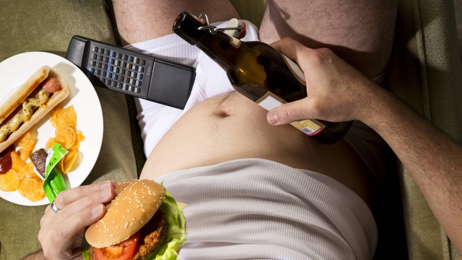 Ein Mann sitzt in der Unterhose auf der Couch mit einer Bierflasche und einem Hamburger in seinen Händen.