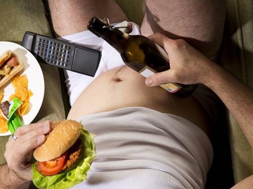 Ein Mann sitzt in der Unterhose auf der Couch mit einer Bierflasche und einem Hamburger in seinen Händen.