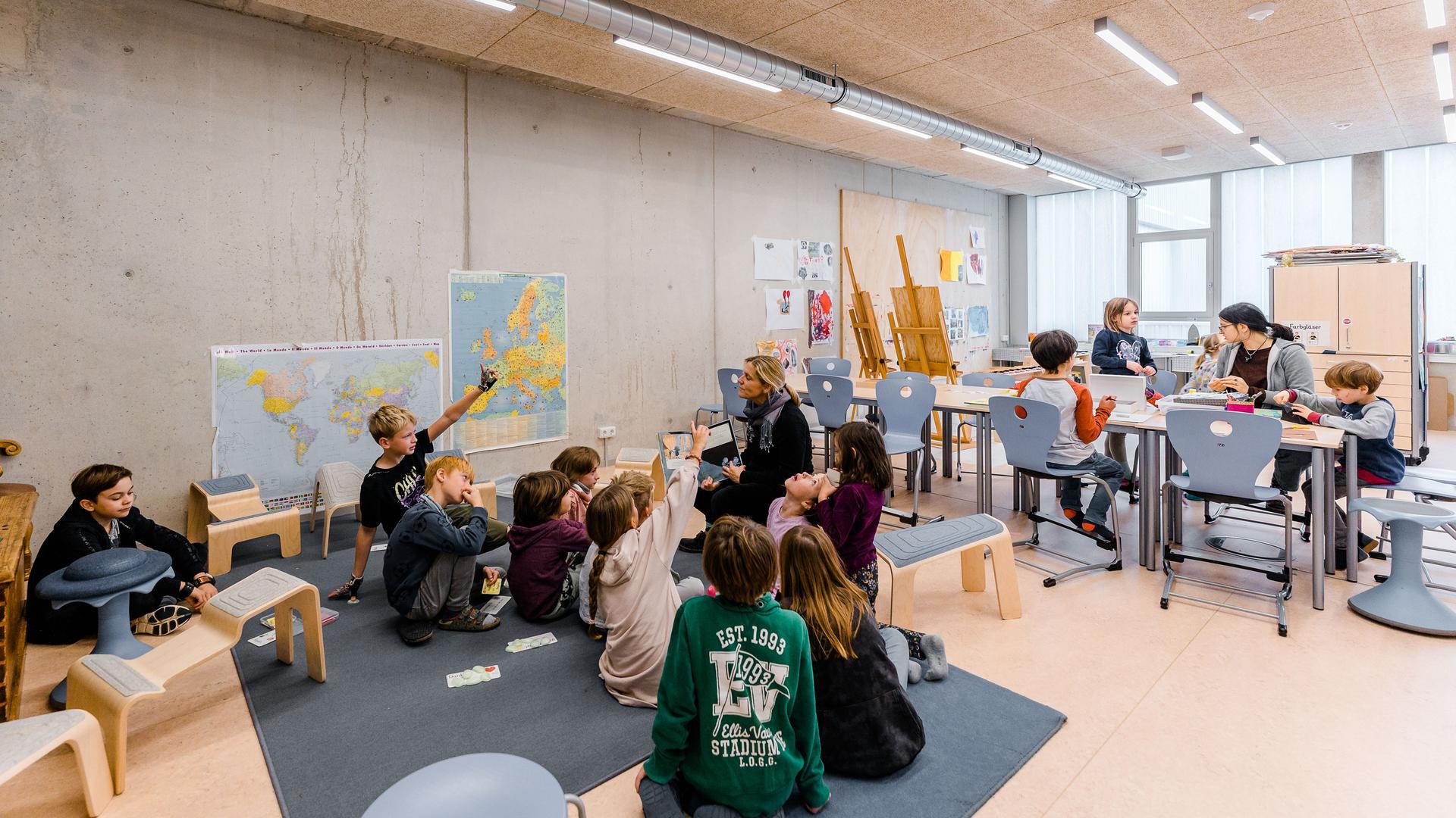 Kinder sitzen um eine Lehrerin herum auf dem Boden eines Klassenzimmers. Im Hintergrund sitzen weitere Kinder an einem Tisch. An den Wänden hängen Atlaskarten und selbstgemalte Bilder, in einer Ecke stehen Staffeleien.