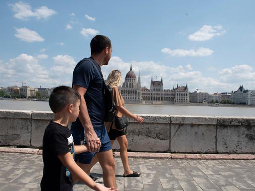 Eine Familie läuft am Ufer der Donau in Budapest entlang, auf der anderen Flussseite ist das Parlamentsgebäude zu sehen