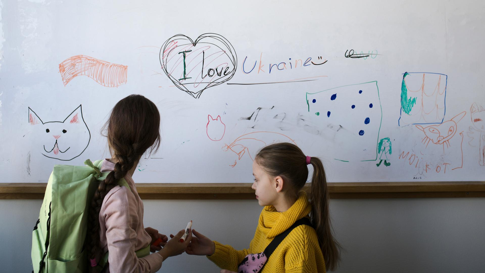 Zwei Mädchen stehen in einem Klassenraum vor einer weißen Tafel, auf der Katzen und ein Herz aufgezeichnet sind, dazu der Satz: "I love Ukraine"