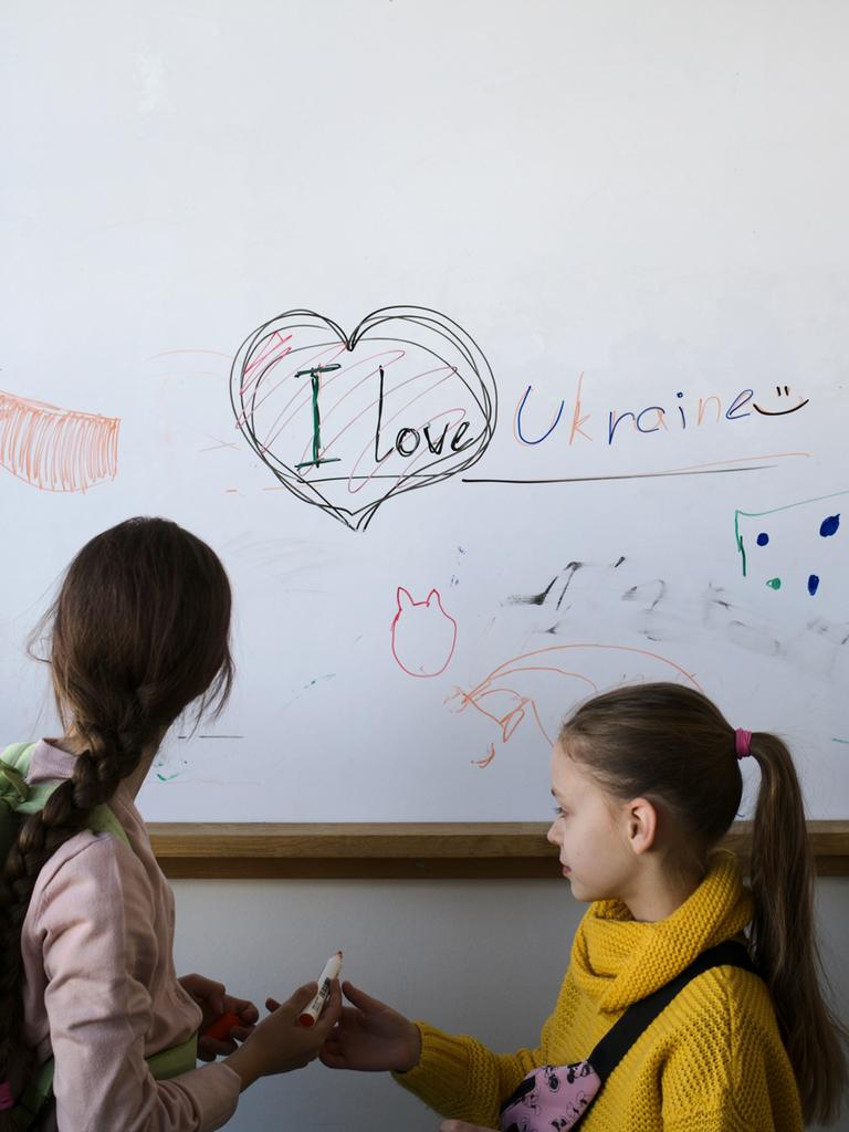 Zwei Mädchen stehen in einem Klassenraum vor einer weißen Tafel, auf der Katzen und ein Herz aufgezeichnet sind, dazu der Satz: "I love Ukraine"