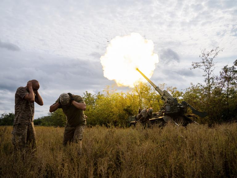 Ukrainische Soldaten feuern an der südlichen Frontlinie der Ukraine am 15. September 2022. Zwei Soldaten gehen in Deckung und halten sich die Ohren zu. Eine Rauchsäule steigt auf.