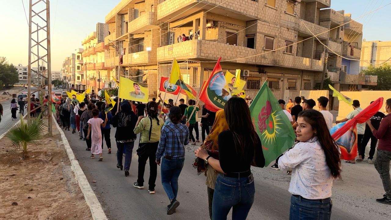 Grüne, gelbe und rote Fahnen schwenken mehrere hundert Menschen und ziehen durch die Straßen der kurdischen Stadt Kobanê. Junge Frauen und Männer und Ältere sind vertreten. Im Hintergrund steht ein Häuserblock.
