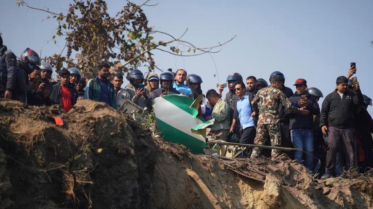 Viele Menschen stehen am Rand eines Trümmerfelds. Vorn im Bild sind die Überreste des abgestürzten Flugzeugs der Yeti Airlines zu sehen. Es steigt Rauch aus den Trümmern auf.