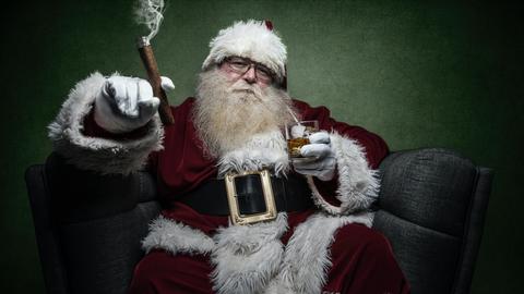 Ein dicker Weihnachtsmann sitzt mit Zigarre und Whiskey entspannt in einem breiten Sessel vor gründer Wand.