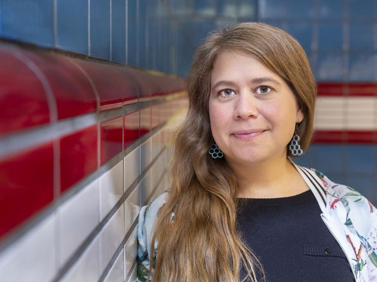 Julia von Lucadou steht angelehnt an eine Fliesenwand, im Hintergrund ist auch eine Fliesenwand. Sie hat lange Haare, die sie mir Seitenscheitel trägt. Ihre Jacke ist hell, ihr T-Shirt dunkelblau.