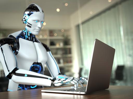 Das Bild zeigt eine 3D-Illustration eines Roboters, der auf einem Laptop schreibt. Der Roboter sitzt in einem Büro an einem Schreibtisch. Es handelt sich nicht um einen realen Roboter sondern um eine Visualisierung. 