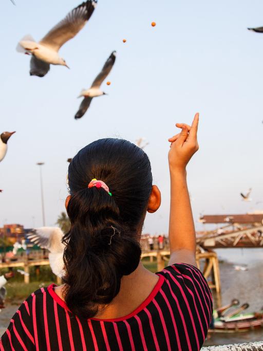 Rear view of women flying birds. Zu sehen: eine Frau mit dem Rücken zur Kamera in einem schwarz-rot längsgestreiften Kleid füttert auf einem Platz Vögel