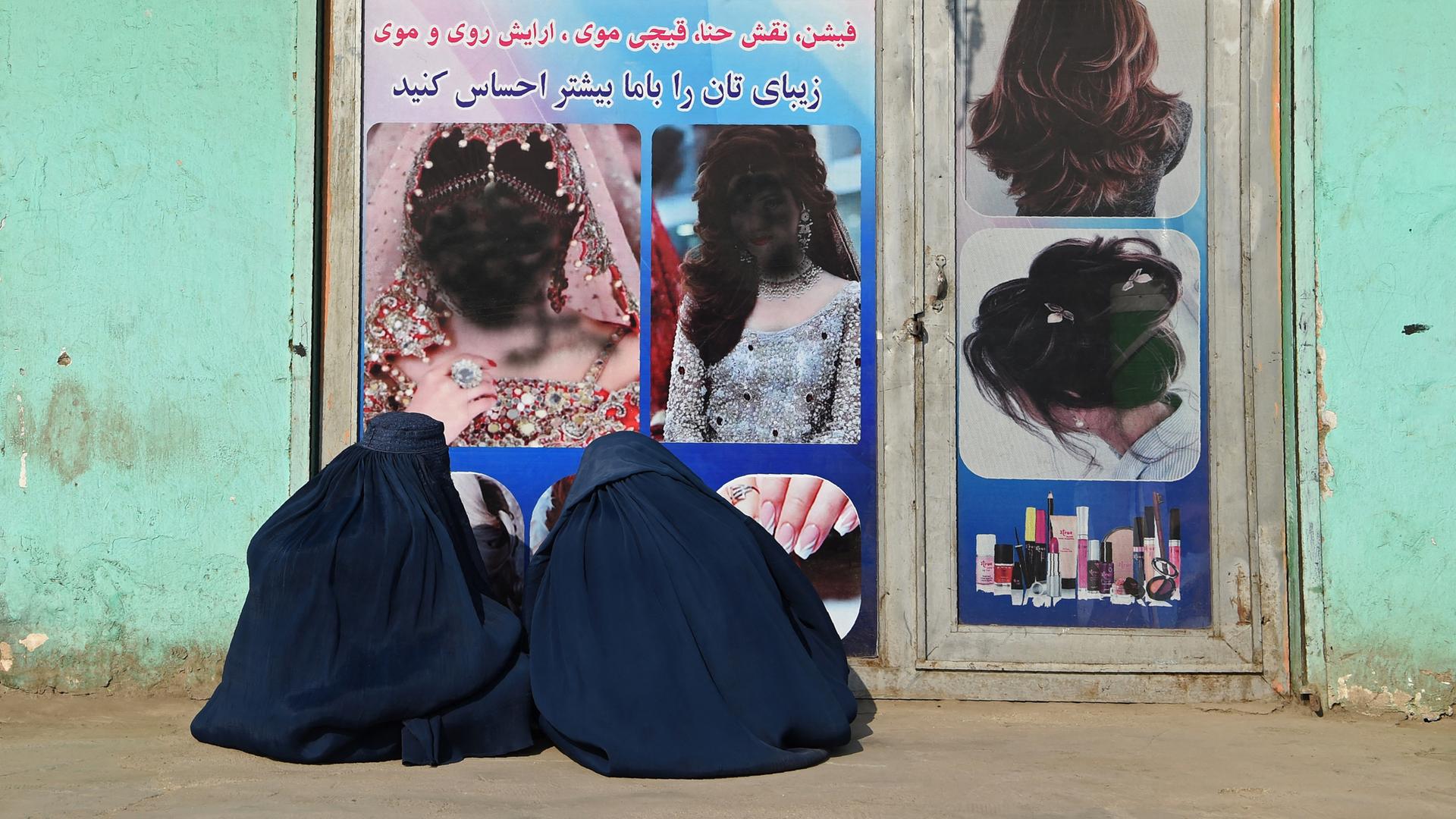 Zwei afghanische Frauen, in Burkas verhüllt, sitzen vor einem geschlossenen Schönheitssalon. Die Frauengesichter im Schaufenster sind mit schwarzer Farbe übersprüht worden. Jalalabad, 13.1. 2021.
