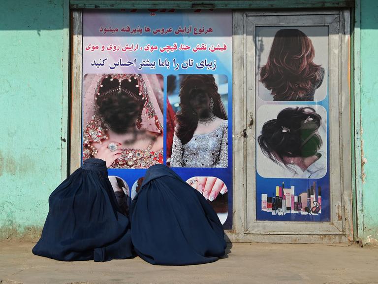 Zwei afghanische Frauen, in Burkas verhüllt, sitzen vor einem geschlossenen Schönheitssalon. Die Frauengesichter im Schaufenster sind mit schwarzer Farbe übersprüht worden. Jalalabad, 13.1. 2021.