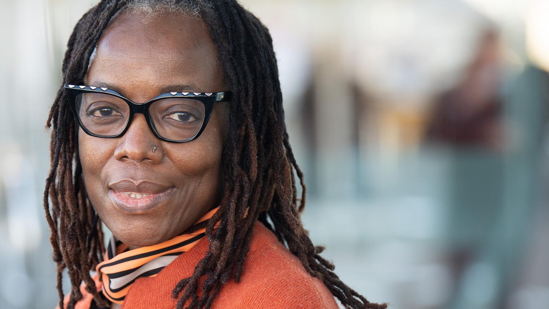 Porträtaufnahme der simbabwischen Autorin Tsitsi Dangarembga. Sie ist Trägerin des Friedenspreises des Deutschen Buchhandels 2021. Dangarembga trägt eine orangefarbene Jacke über einer orange-braun-weiß gemusterten Bluse. Ihre Haare trägt sie zu vielen Zöpfen geflochten. Porträtaufnahme der simbabwischen Autorin Tsitsi Dangarembga. Sie ist Trägerin des Friedenspreises des Deutschen Buchhandels 2021. Dangarembga trägt eine orangefarbene Jacke über einer orange-braun-weiß gemusterten Bluse. Ihre Haare trägt sie zu vielen Zöpfen geflochten.