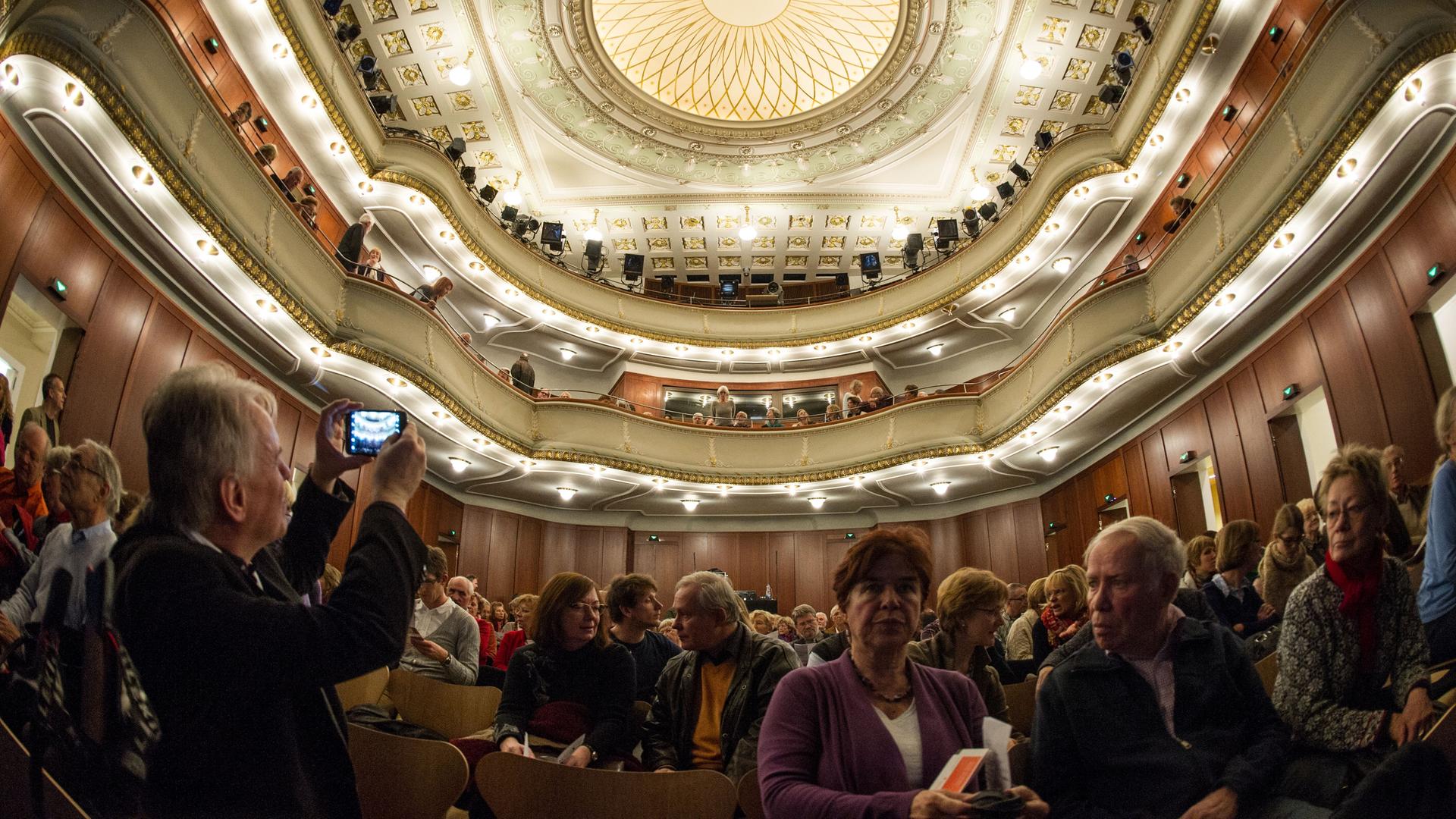 Zuschauer sitzen im Alten Saal des Theaters Heidelberg. Ein Mann macht ein Foto der mit Ornamenten verzierten Decke. Zuschauer sitzen im Alten Saal des Theaters Heidelberg. Ein Mann macht ein Foto der mit Ornamenten verzierten Decke.