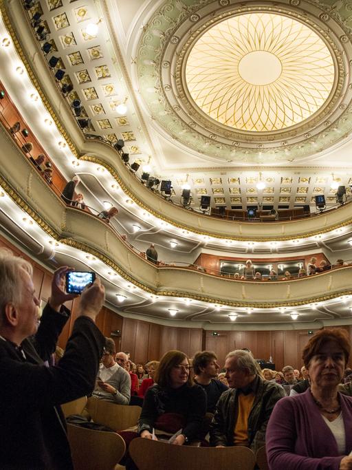 Zuschauer sitzen im Alten Saal des Theaters Heidelberg. Ein Mann macht ein Foto der mit Ornamenten verzierten Decke.
