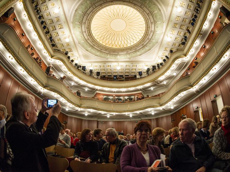 Zuschauer sitzen im Alten Saal des Theaters Heidelberg. Ein Mann macht ein Foto der mit Ornamenten verzierten Decke.