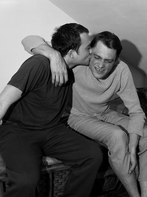 Schwarz-weiß Foto von einem Brüderpaar, das sich, auf einem Bett sitzend, in einer Dachkammer umarmt. Der eine Bruder küsst den anderen mit Begeisterung auf den Hals, der scheint nicht so angetan.