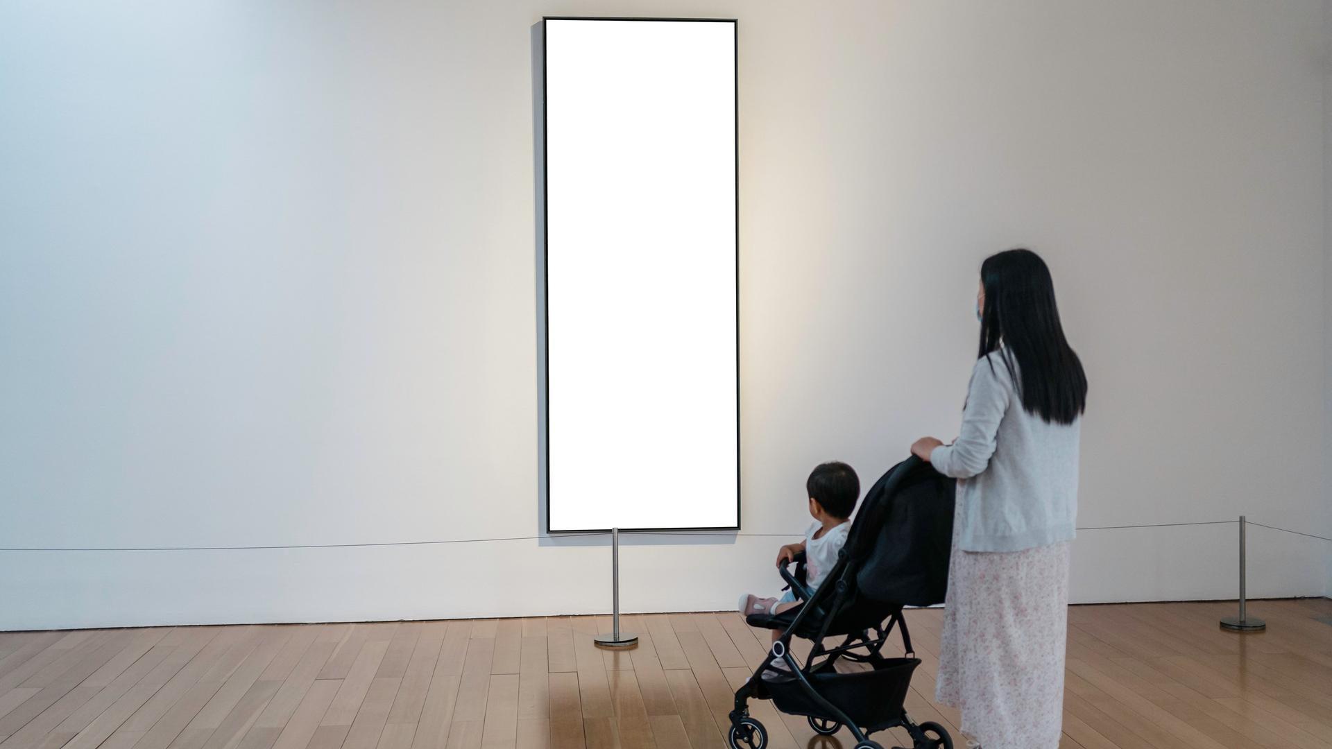 Eine Frau mit einem Kinderwagen in einer Galerie vor einem leeren weißen Bild.